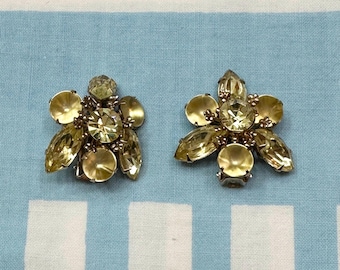 Pode incluir: Um par de brincos em tom dourado com um aglomerado de strass amarelos transparentes em forma de flor.