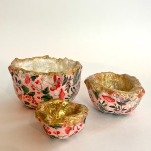 May include: Three decorative bowls with a pink floral pattern featuring red berries and green leaves. The rims and interiors of the bowls are gold. The bowls vary in size, with the largest bowl on the left and the smallest in the center.