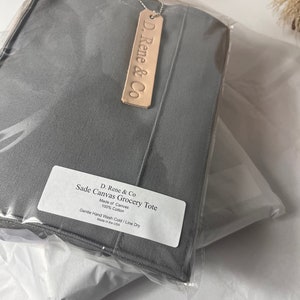 May include: A gray canvas grocery tote bag with a leather tag that says "D. Rene & Co". The tote bag is made of 100% cotton and has the text "Sade Canvas Grocery Tote" and "Gentle Hand Wash Cold / Line Dry" printed on a white label.