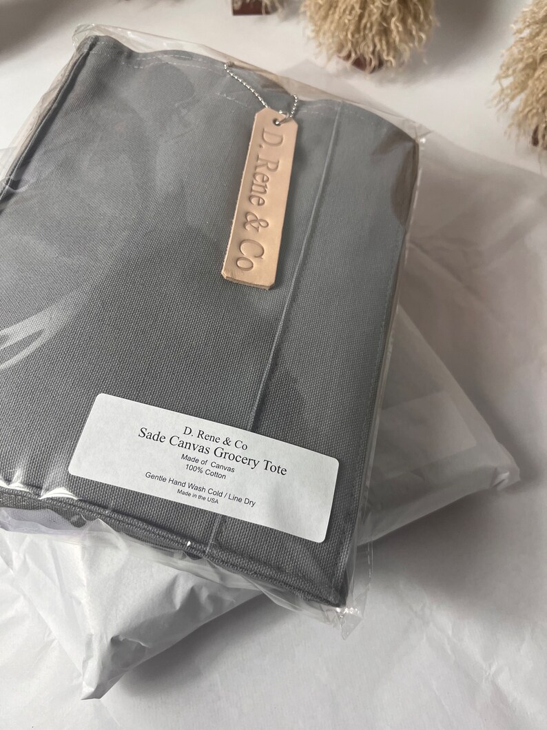 May include: A gray canvas grocery tote bag with a leather tag that says "D. Rene & Co". The tote bag is made of 100% cotton and has the text "Sade Canvas Grocery Tote" and "Gentle Hand Wash Cold / Line Dry" printed on a white label.