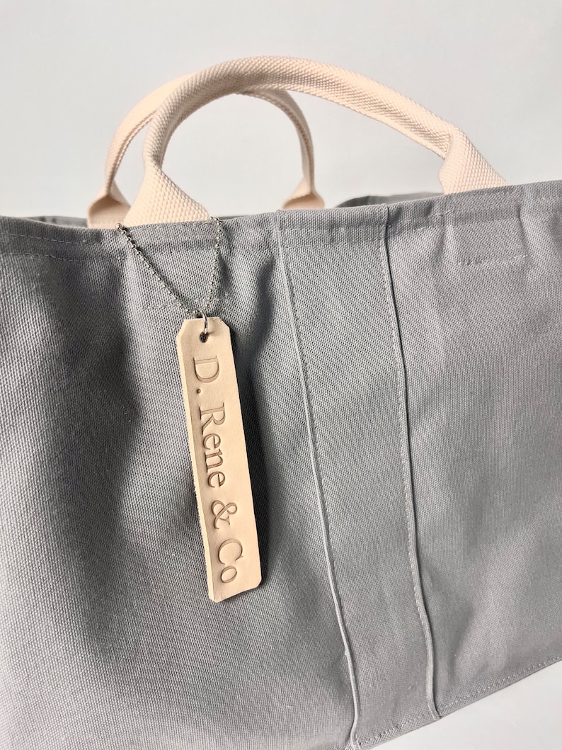 May include: A gray canvas tote bag with cream colored handles and a leather tag that says "D. Rene & Co."