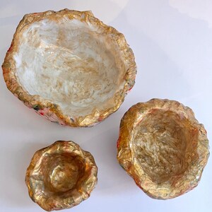 May include: Three decorative bowls with irregular edges. The bowls are in varying sizes, with a gold-colored rim and interior. The interior of the bowls has a marbled effect with white and gold tones. The exterior has a floral pattern.