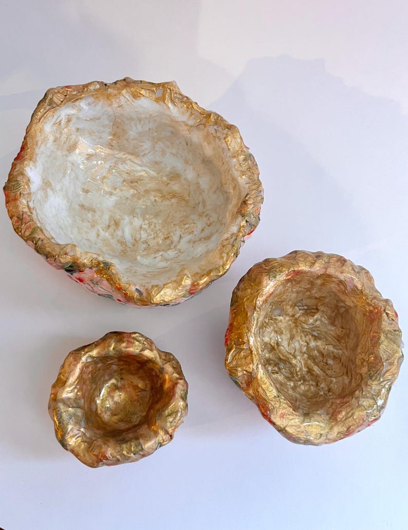May include: Three decorative bowls with irregular edges. The bowls are in varying sizes, with a gold-colored rim and interior. The interior of the bowls has a marbled effect with white and gold tones. The exterior has a floral pattern.