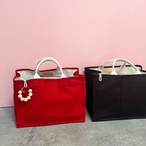 May include: Two tote bags, one red and one black, with white handles. The red bag has a wooden bead charm attached to it.