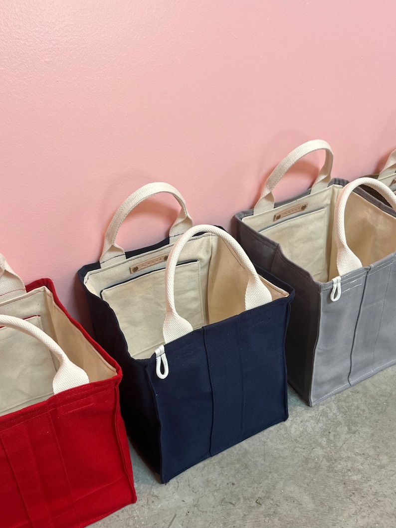 May include: Four tote bags in different colors: red, navy blue, gray, and white. The bags have white handles and a zipper closure.