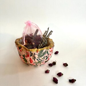 May include: A decorative pink bowl with a floral pattern and gold trim, containing a pink mesh bag, dried lavender, and rose buds. The bowl is surrounded by scattered rose buds, creating a natural, botanical theme.