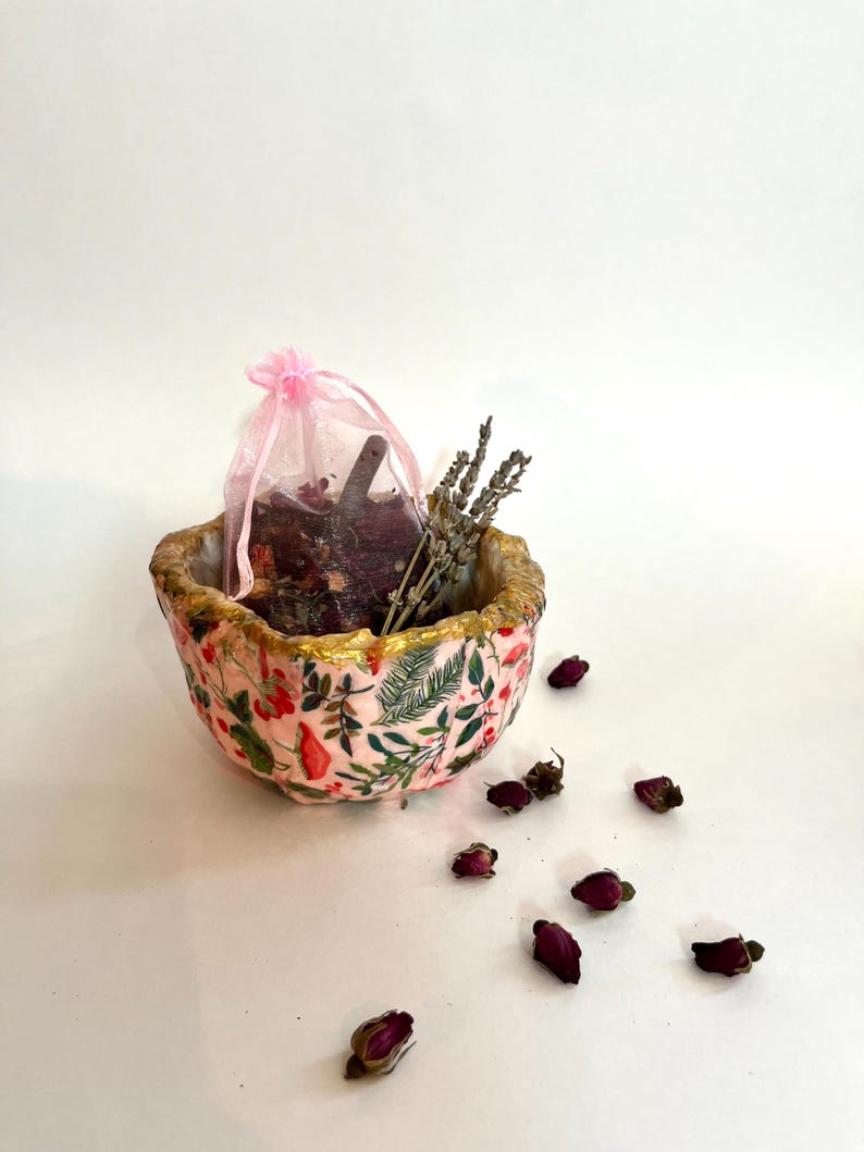 May include: A decorative pink bowl with a floral pattern and gold trim, containing a pink mesh bag, dried lavender, and rose buds. The bowl is surrounded by scattered rose buds, creating a natural, botanical theme.