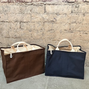 May include: Two large canvas tote bags, one brown and one navy blue, with white handles. The bags are sitting on a concrete surface in front of a brick wall.
