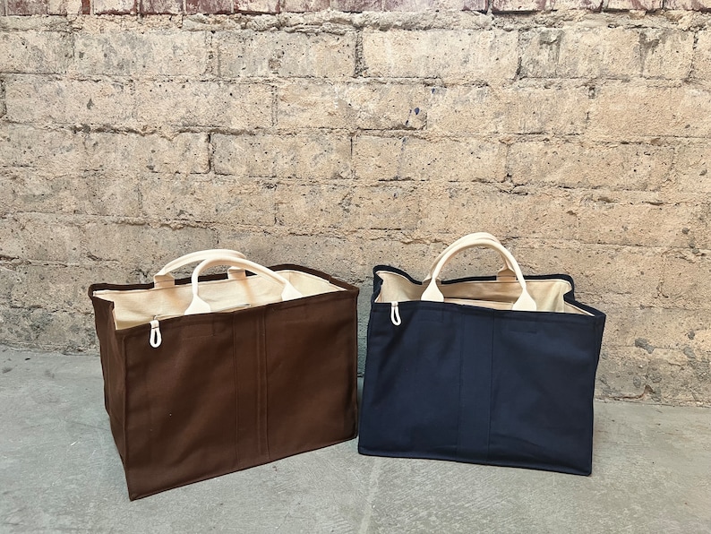 May include: Two large canvas tote bags, one brown and one navy blue, with white handles. The bags are sitting on a concrete surface in front of a brick wall.