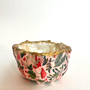 May include: A small, decorative bowl with a pink floral pattern. The bowl's interior is white, and the rim is trimmed with gold. The floral design includes red berries, green leaves, and other botanical elements.