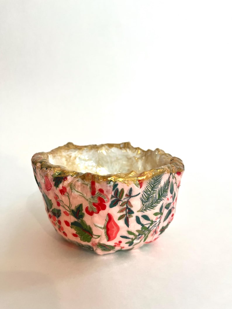 May include: A small, decorative bowl with a pink floral pattern. The bowl's interior is white, and the rim is trimmed with gold. The floral design includes red berries, green leaves, and other botanical elements.