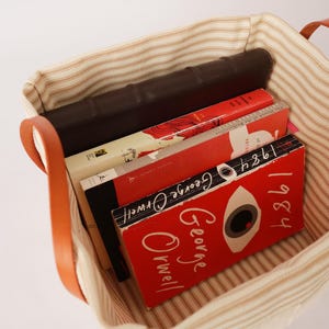 May include: A canvas tote bag with brown leather handles, filled with books. The books have red and white covers, including one with the title "1984" by George Orwell. The bag has a striped pattern.