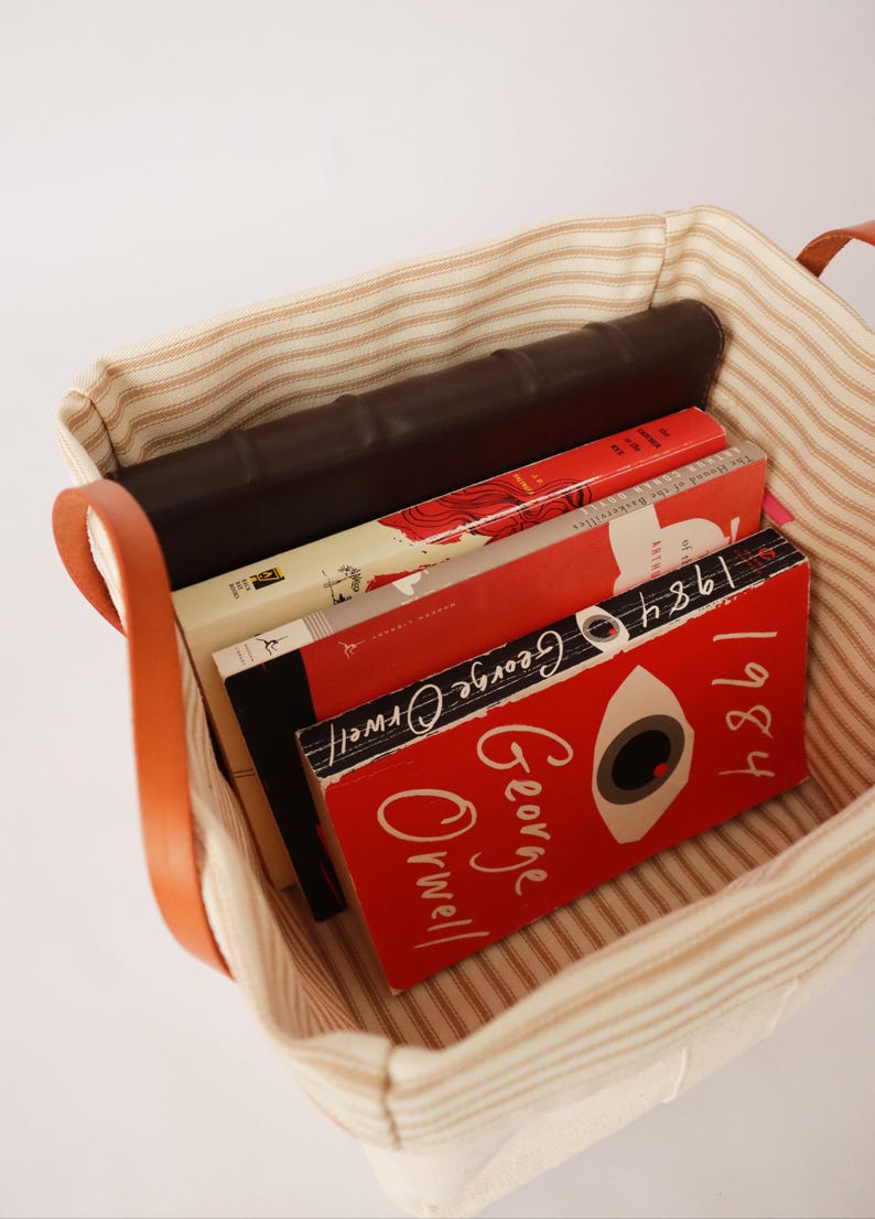 May include: A canvas tote bag with brown leather handles, filled with books. The books have red and white covers, including one with the title "1984" by George Orwell. The bag has a striped pattern.