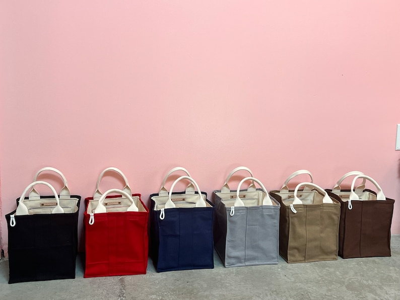 May include: Seven canvas tote bags in different colors, including black, red, navy blue, gray, beige, and brown. The bags have white handles and are lined with white fabric.