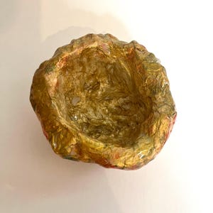 May include: A handmade, irregularly shaped bowl with a textured surface. The interior is a shimmering gold, while the exterior features a mix of gold, red, and green hues. The bowl is set against a white background.