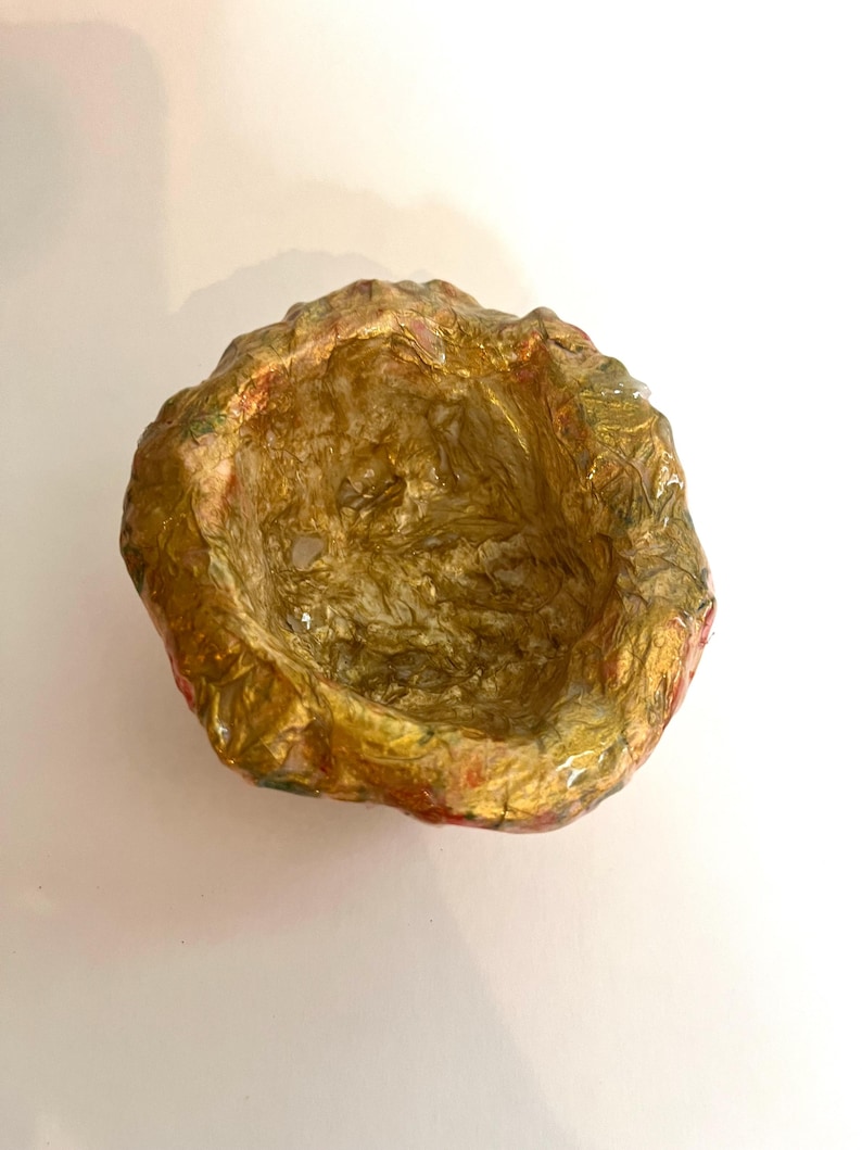 May include: A handmade, irregularly shaped bowl with a textured surface. The interior is a shimmering gold, while the exterior features a mix of gold, red, and green hues. The bowl is set against a white background.