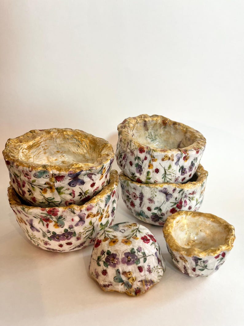 Pode incluir: Um conjunto de seis tigelas decorativas com um padr&atilde;o floral e de borboletas. As tigelas apresentam flores vermelhas, roxas e amarelas, com bordas douradas. As tigelas s&atilde;o de tamanhos e formas variadas, com uma textura.