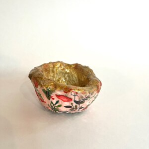 May include: A small, handmade bowl with a pink floral pattern and gold interior. The bowl's exterior features a delicate design of red flowers and green leaves. The rim is textured and painted gold, adding a touch of elegance to this decorative piece.