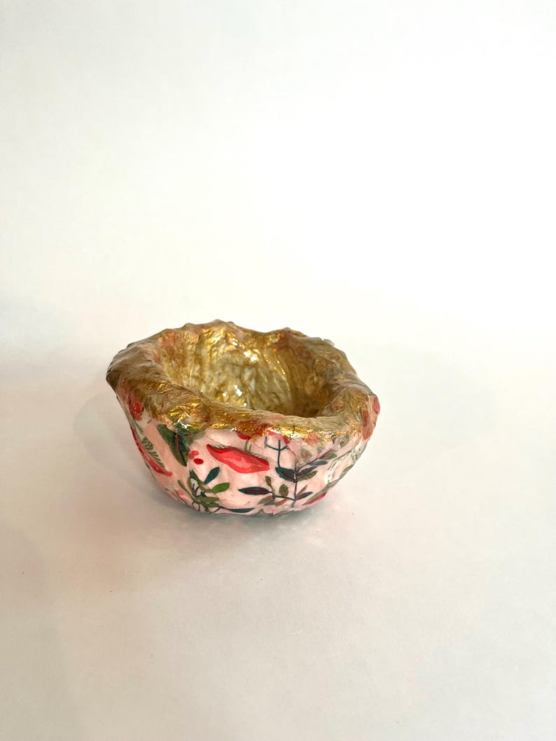 May include: A small, handmade bowl with a pink floral pattern and gold interior. The bowl's exterior features a delicate design of red flowers and green leaves. The rim is textured and painted gold, adding a touch of elegance to this decorative piece.