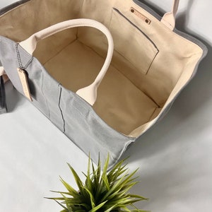 May include: A gray canvas tote bag with a cream-colored lining and a small pocket on the inside. The bag has two cream-colored handles and a leather tag with the word "MADEWELL" on it.