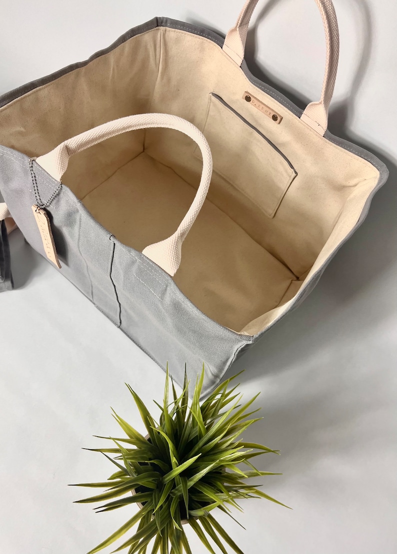 May include: A gray canvas tote bag with a cream-colored lining and a small pocket on the inside. The bag has two cream-colored handles and a leather tag with the word "MADEWELL" on it.