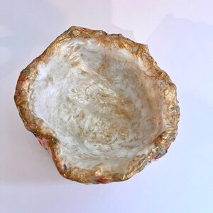May include: A handmade, decorative bowl with a textured, irregular rim. The interior is white with gold accents, and the rim features gold and floral patterns. The bowl is designed to hold small items.