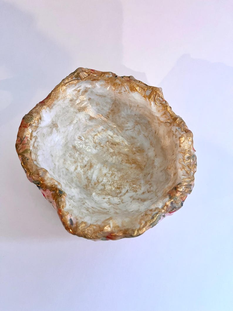 May include: A handmade, decorative bowl with a textured, irregular rim. The interior is white with gold accents, and the rim features gold and floral patterns. The bowl is designed to hold small items.