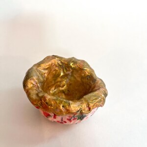 May include: A small, handmade bowl with a textured, gold-colored interior and rim. The exterior is pink with a floral pattern. The bowl is likely used for decorative purposes or holding small items.