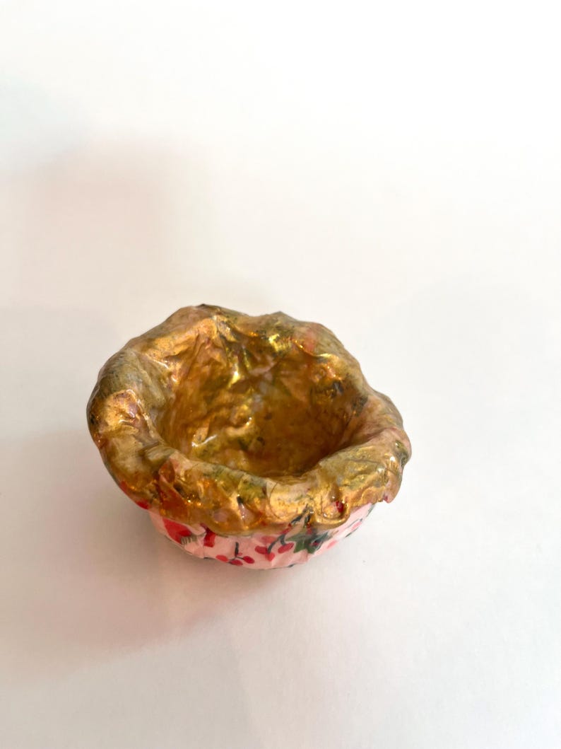 May include: A small, handmade bowl with a textured, gold-colored interior and rim. The exterior is pink with a floral pattern. The bowl is likely used for decorative purposes or holding small items.