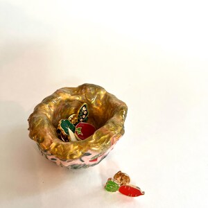 May include: A small, handmade ceramic bowl with a pink and gold exterior. The bowl contains several enamel pins, including a tomato, a leaf, and a carrot. The bowl is sitting on a white surface.