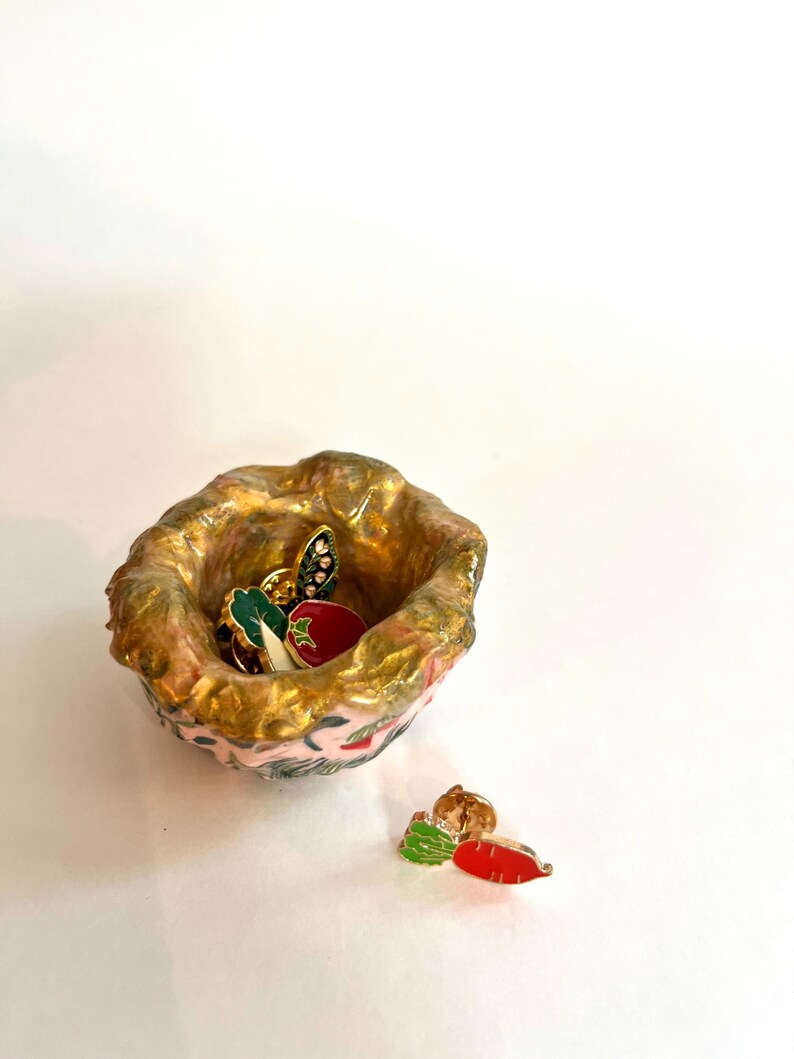 May include: A small, handmade ceramic bowl with a pink and gold exterior. The bowl contains several enamel pins, including a tomato, a leaf, and a carrot. The bowl is sitting on a white surface.