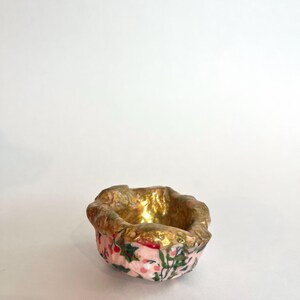 May include: A small, decorative ceramic bowl with a pink exterior featuring a floral pattern of red berries and green leaves. The interior and rim are gold-colored, creating a contrast. The bowl is set against a white background.