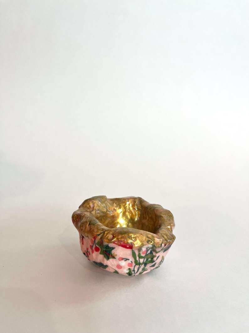 May include: A small, decorative ceramic bowl with a pink exterior featuring a floral pattern of red berries and green leaves. The interior and rim are gold-colored, creating a contrast. The bowl is set against a white background.