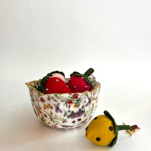 Pode incluir: Uma tigela decorativa cheia de dois ornamentos vermelhos em forma de morango com caules verdes e detalhes prateados. Um ornamento amarelo em forma de lim&atilde;o com pontos pretos e um caule verde est&aacute; por perto.
