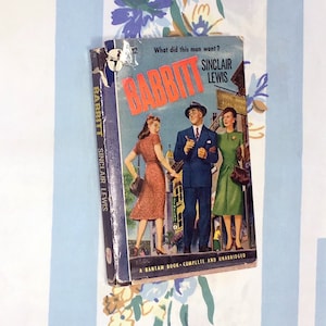May include: A vintage paperback book cover with the title "Babbitt" by Sinclair Lewis. The cover features a colorful illustration of three people walking down a street. The text "What did this man want?" is printed at the top of the cover.