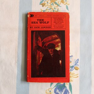 May include: A red paperback book with the title "The Sea Wolf" by Jack London. The cover features a black and white illustration of a man in a dark coat and hat standing in a doorway.