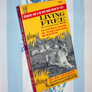 May include: A yellow paperback book titled "Living Free" by Joy Adamson. The cover features a photo of a lioness and her cubs in a grassy field. The book is a sequel to "Born Free".