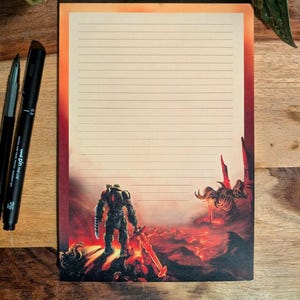 May include: Lined notepad with a vibrant illustration of a futuristic warrior standing on a fiery landscape. The notepad has a gradient border and is accompanied by two black pens.