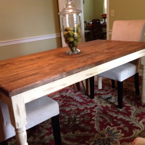 May include: A rectangular wooden dining table with a dark stained top and distressed white legs. Two upholstered chairs with light gray fabric are positioned on either side. A glass jar filled with green fruit sits on the table.