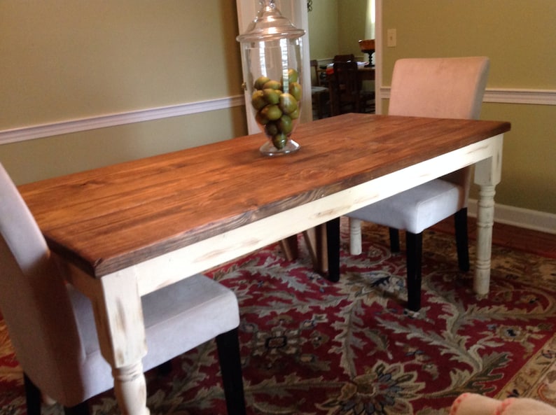 May include: A rectangular wooden dining table with a dark stained top and distressed white legs. Two upholstered chairs with light gray fabric are positioned on either side. A glass jar filled with green fruit sits on the table.