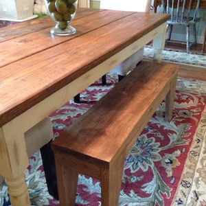 May include: A rustic wooden dining table with a matching bench. The table has a dark stained top and cream-colored legs. The bench is made of the same wood and stain. The table is approximately 72 inches long.
