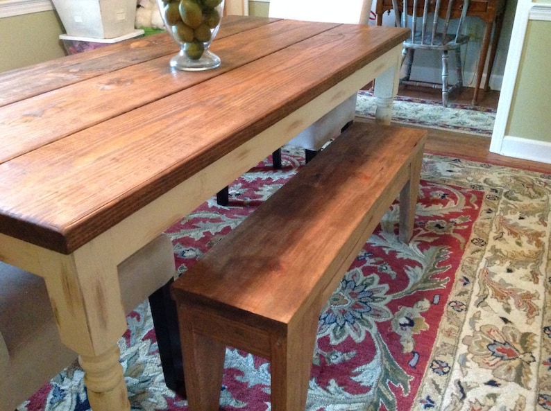 May include: A rustic wooden dining table with a matching bench. The table has a dark stained top and cream-colored legs. The bench is made of the same wood and stain. The table is approximately 72 inches long.