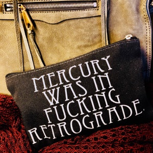 Könnte beinhalten: Schwarze Kosmetiktasche aus Canvas mit weißem Text, der "Mercury was in fucking retrograde" lautet.