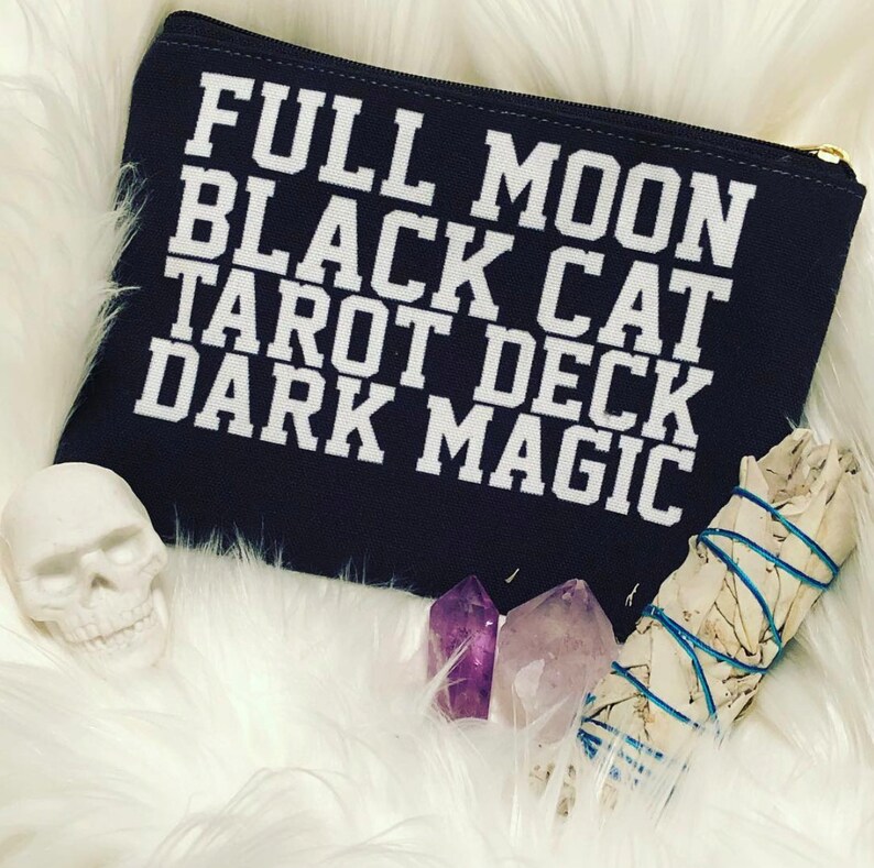 K&ouml;nnte beinhalten: Eine dunkelblaue Stofftasche mit wei&szlig;em Text, der "FULL MOON BLACK CAT TAROT DECK DARK MAGIC" lautet. Die Tasche ist von wei&szlig;em Kunstfell, einem wei&szlig;en Sch&auml;del, zwei Amethystkristallen und einem wei&szlig;en Salbeistab umgeben.