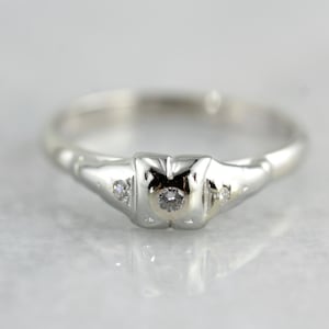 Puede incluir: Un anillo de plata con un pequeño diamante redondo en el centro y dos diamantes más pequeños a cada lado.