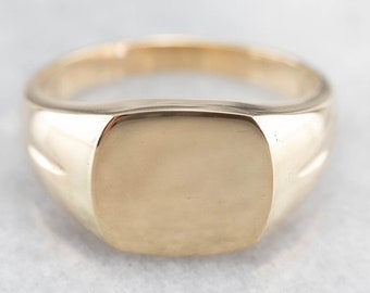 May include: A gold signet ring with a smooth, rectangular top. The ring has a slightly rounded band.