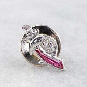 May include: A silver-toned lapel pin featuring a stylized design with a central diamond and a rectangular, vibrant pink gemstone. The pin has intricate detailing and a classic aesthetic, suitable for formal wear or adding a touch of elegance.