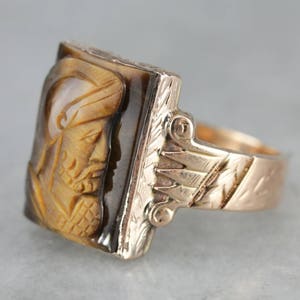 May include: A gold ring with a carved tiger's eye stone featuring a detailed profile of a person. The band is engraved with a floral design.