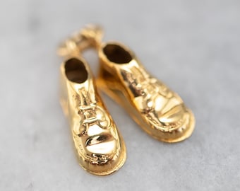Op de afbeelding: Twee gouden schoenbedels in de vorm van een paar schoenen. De schoenen hebben veters en een afgeronde neus.