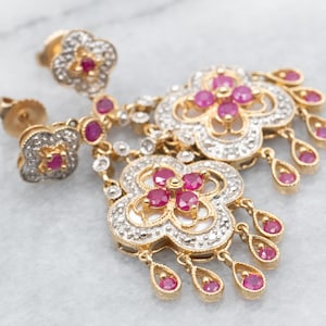 以下が含まれることがあります： 花柄のデザインのゴールドのドロップイヤリングのペア。イヤリングはピンクの宝石とクリアな石で飾られています